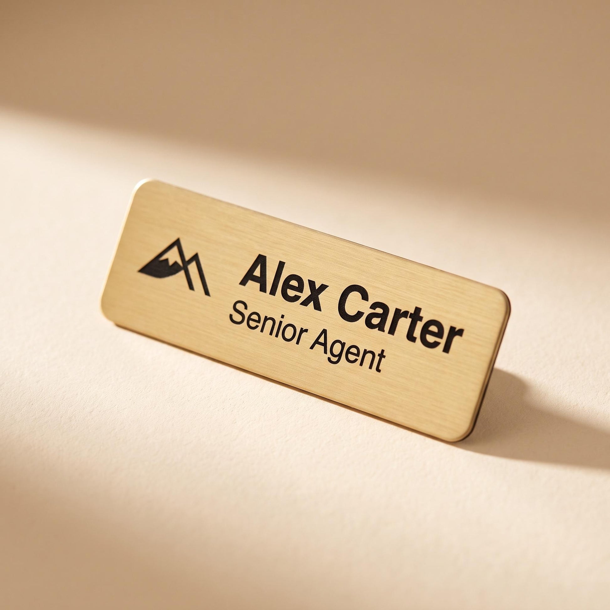 Rectangular brushed-gold custom name badge engraved with a mountain logo, name, and job title, resting on a cream surface.