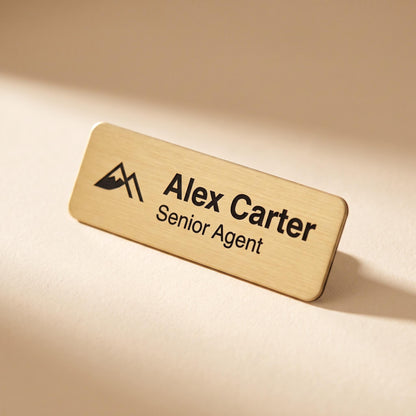 Rectangular brushed-gold custom name badge engraved with a mountain logo, name, and job title, resting on a cream surface.