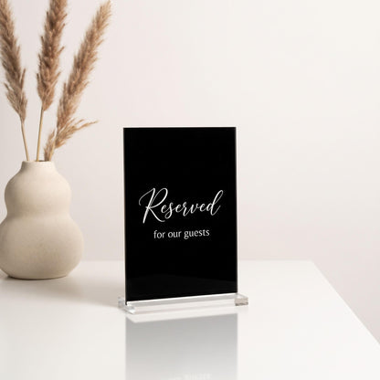Black acrylic Reserved table sign in a clear acrylic block stand on a white tabletop next to a dried pampas grass vase.