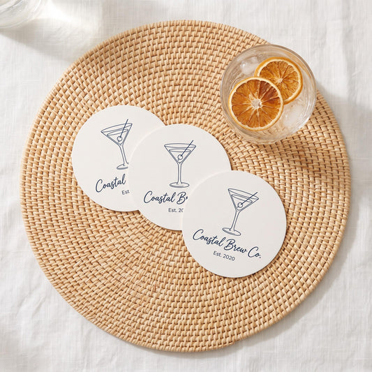 Three round white coasters printed with a navy martini illustration and custom Coastal Brew Co. logo text, arranged on a woven rattan placemat next to a citrus garnished drink.
