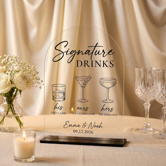 Clear arch-topped acrylic Signature Drinks sign with custom cocktail illustrations and couple names, displayed on a black stand at a wedding reception table.