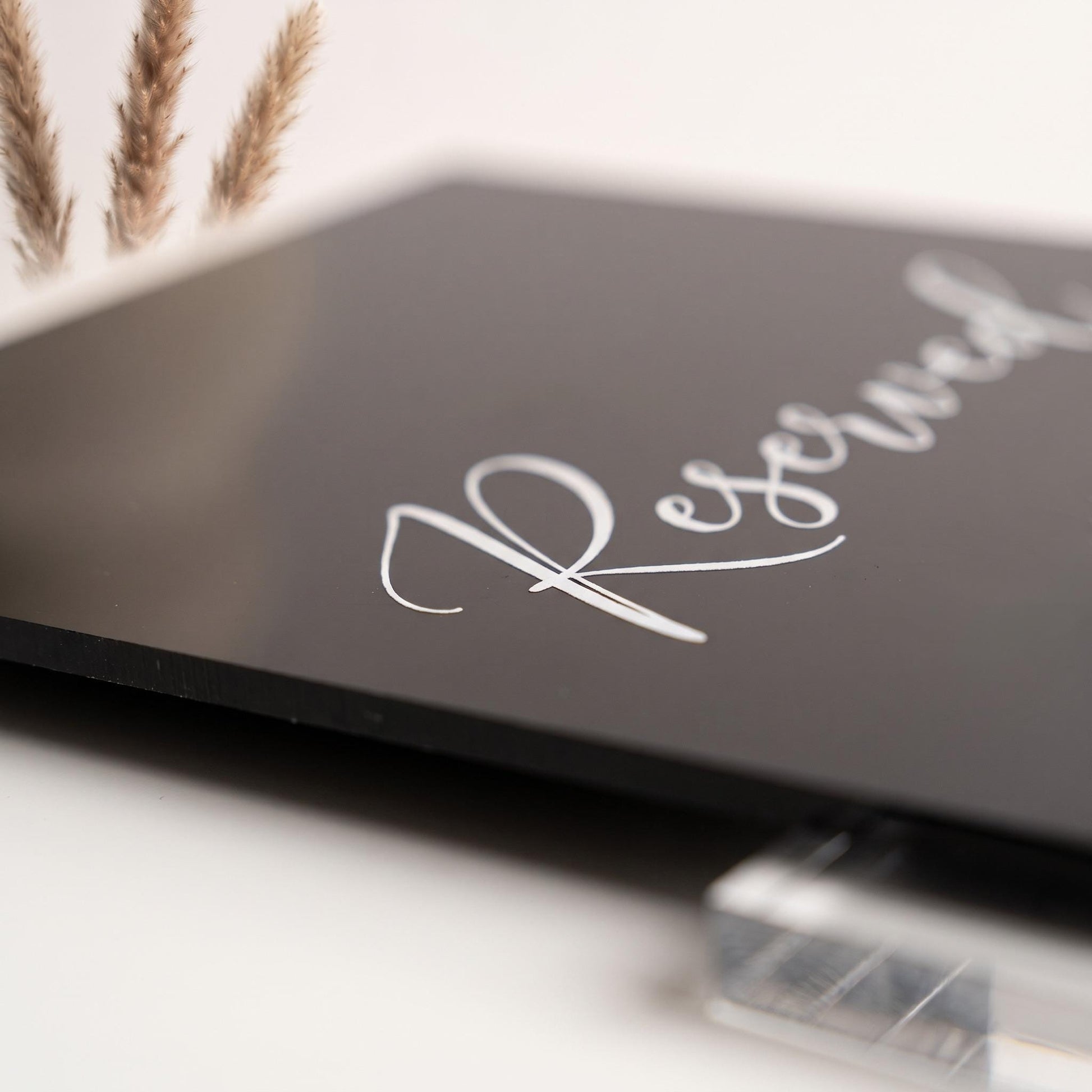 Close-up of the black acrylic table sign showing the white cursive text and clear stand joint.