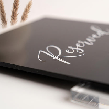 Close-up of the black acrylic table sign showing the white cursive text and clear stand joint.
